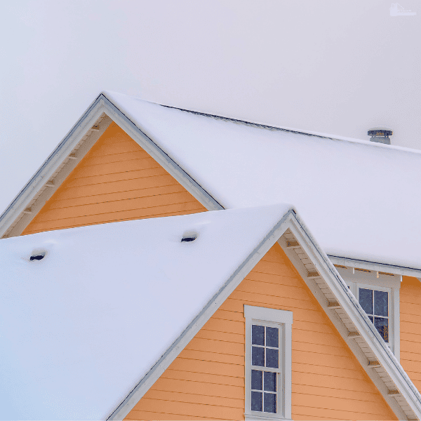 Snow blankets a yellow house’s roof, highlighting an area to inspect for exterior warning signs before winter.