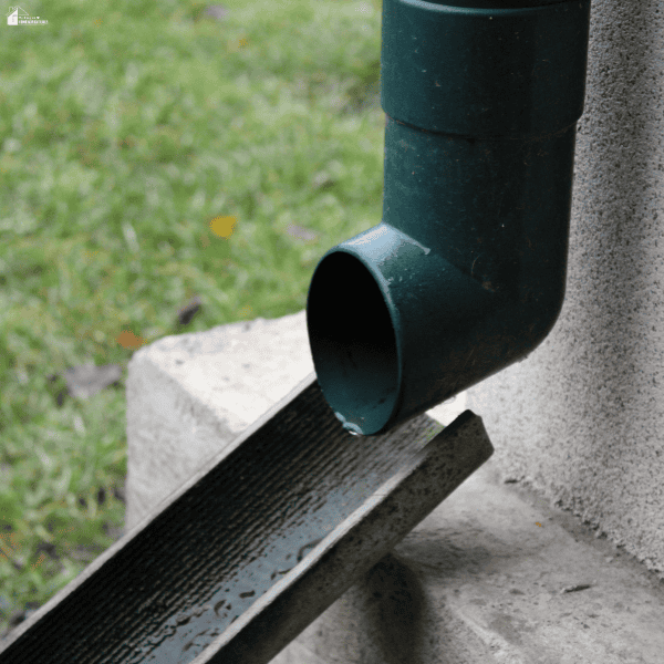 A downspout beside a small home directs rainwater into an angled drainage channel away from the foundation.