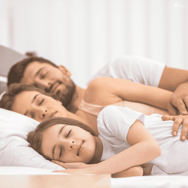 Two adults and a young daughter sleep close together on white bedding, illustrating family rest alongside a bedtime shift.