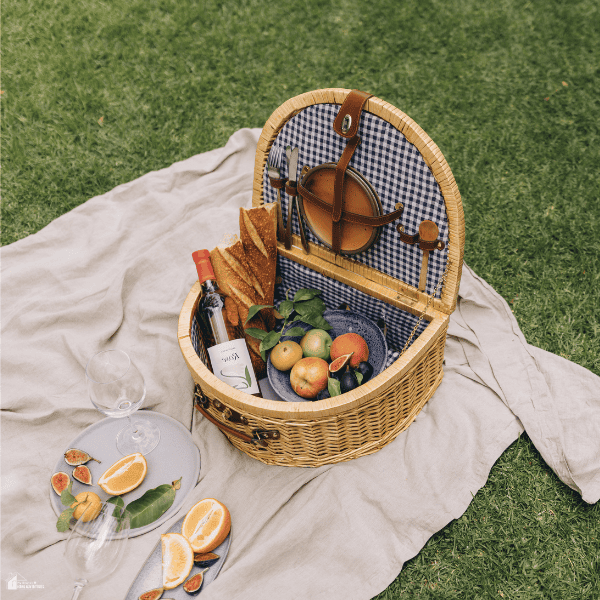 A wicker basket filled with bread and fruit rests on a cream picnic rug arranged for comfortable outdoor lounging.