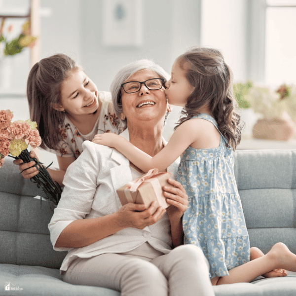 An older woman receives a gift and a kiss from a young girl while an adult woman holds flowers beside their family home’s sofa.