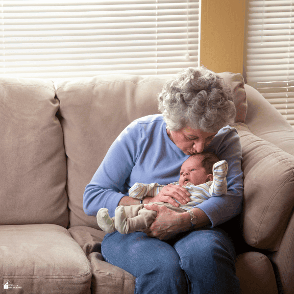 An older woman cradles a newborn and kisses the baby’s forehead while seated on a beige sofa in a family home.