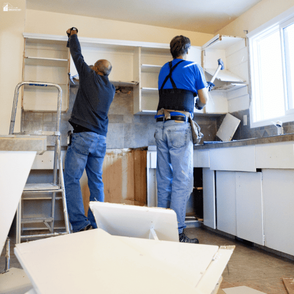Two workers remove upper cabinets during a kitchen renovation, with detached panels and a ladder nearby.